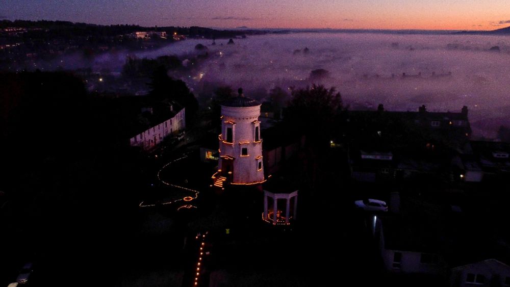 A town of white buildings covered in fairy lights. There is mist coming over the hills