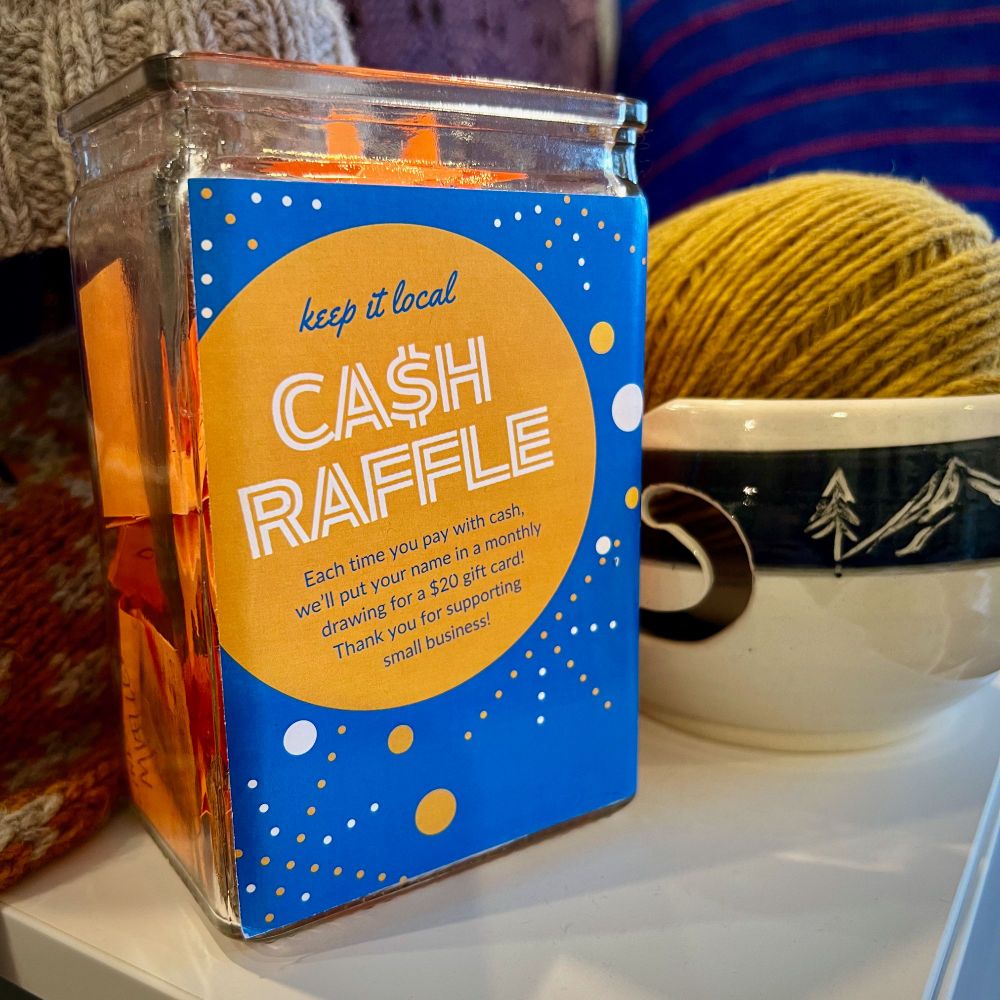 photo of Cash Raffle entry jar at Fancywork Yarn Shop. A ball of mustard colored Wooldreamers Mota yarn is in a ceramic yarn bowl next to the cash raffle jar. 