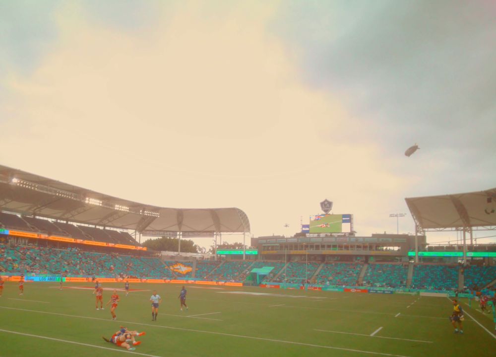 stadium on a cloudy day, a rugby game is happening. There's a pair of women in a tackle in the foreground. 