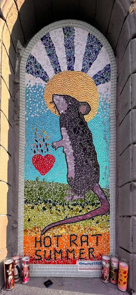 A photograph of the Hot Rat Summer mosaic in Seattle. A rat stands on its hind legs in the grass, head encircled by the sun with black and white rays shooting into the sky.