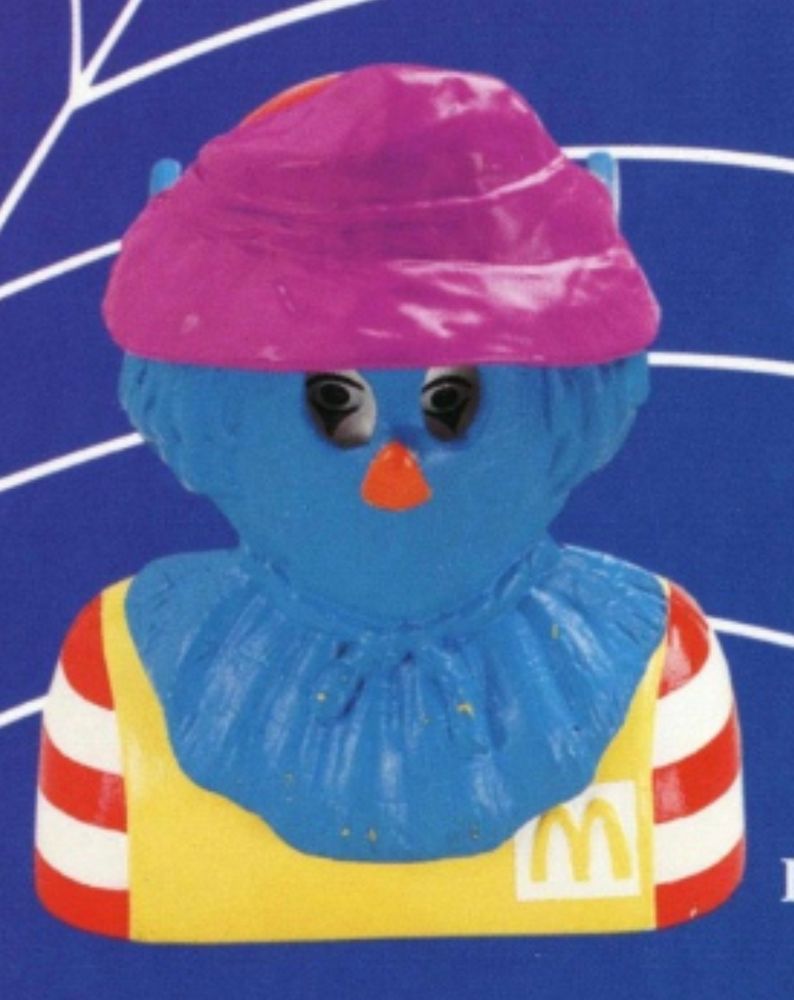 Hard for alt-text to do this justice, but it's a plastic Ronald McDonald (already a clown!) bust wearing a hard plastic, blue and purple scarecrow mask and doily. Ronald's piercing eyes lie in sinister shadow beneath the mask's eye holes.