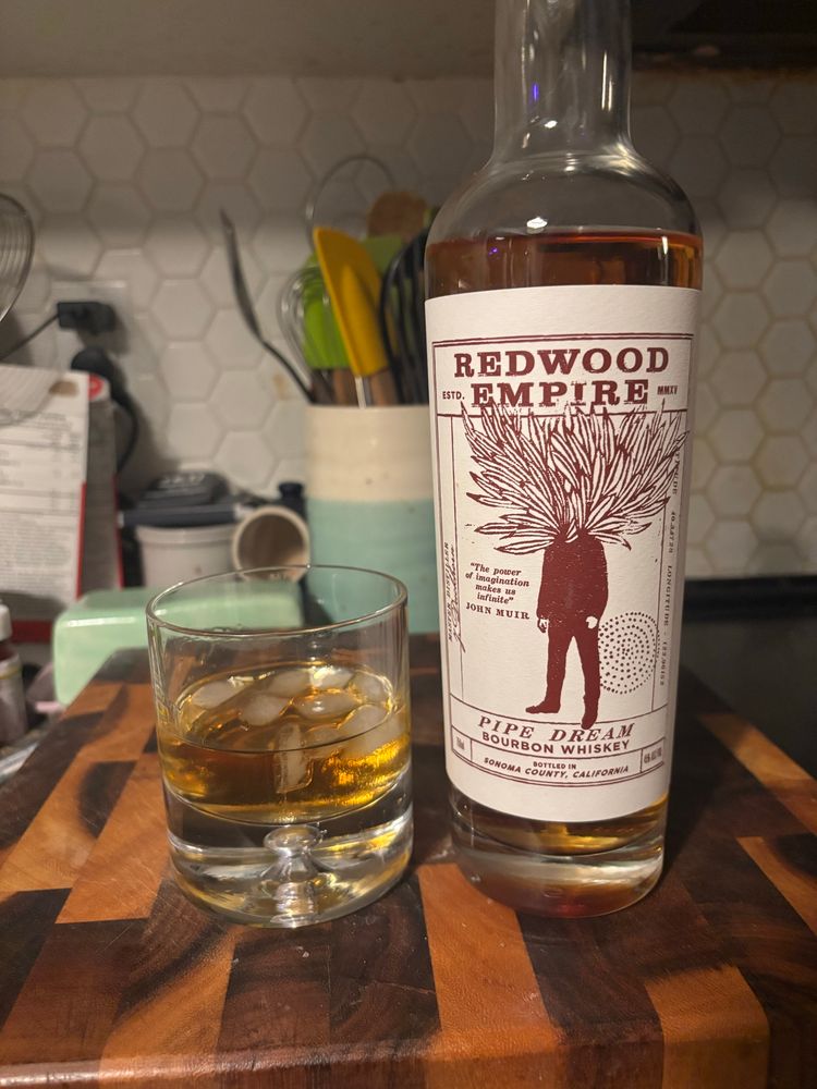 A glass of bourbon with ice, next to a bottle of redwood empire’s pipe dream bourbon