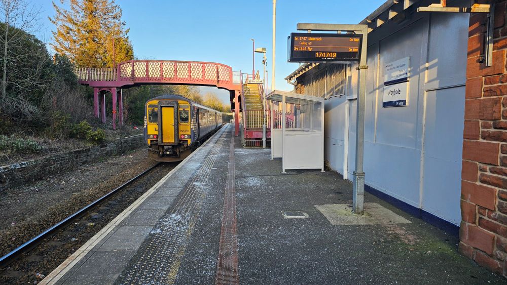 A Class 156 departs Maybole station in 2025. The sun is setting as it departs.