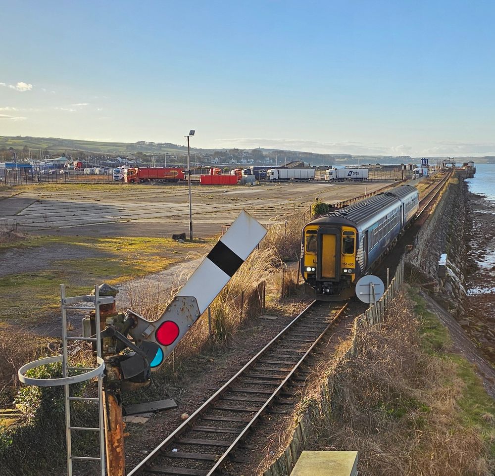 A Class 156 departs for Ayr. The old ferry port lies derelict behind the train