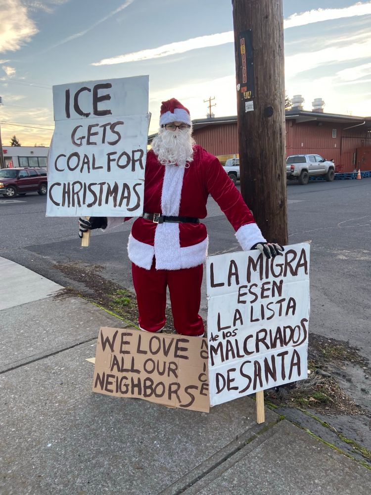 Santa standing on a street corner in an Oregon town with signs that read “ICE gets coal for Christmas,” “La migra esen La lista malcrados de Santa,” and “We Love All Our Neighbors.”