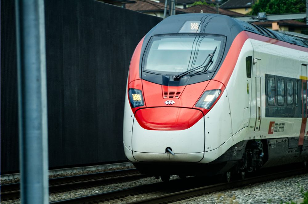Ein SBB Giruno Schnellzug im Tessin fährt an einer grauen Lärmschutzwand vorbei.