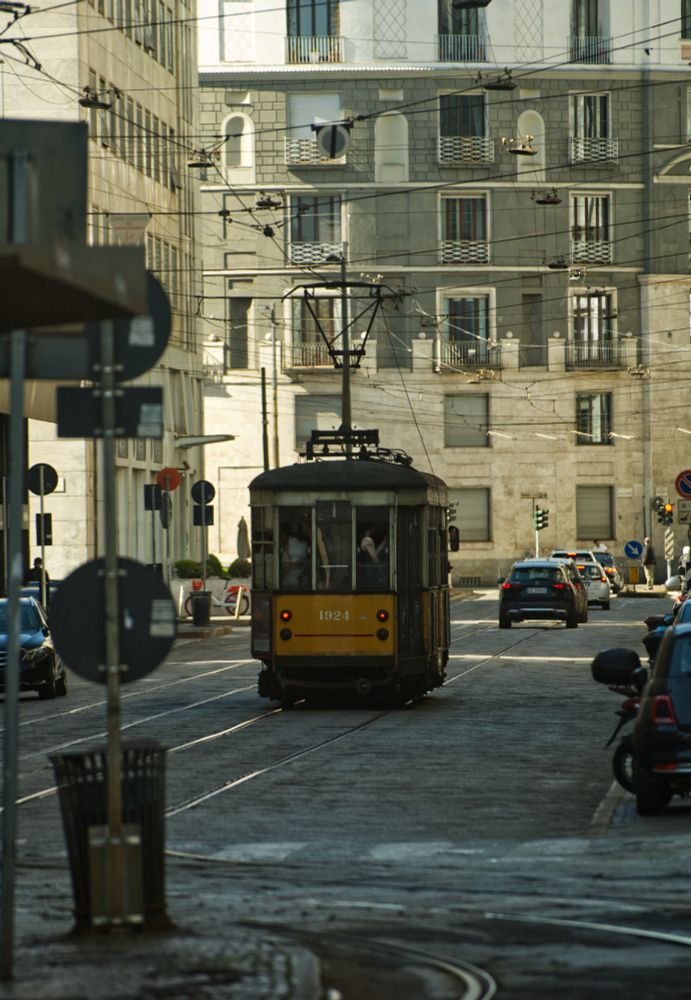 Eine historische Strassenbahn fährt in einer Mailänder Strasse davon. Die Strasse ist im Schatten und im Hintergrund sind die Fassaden der Häuser im Sonnenlicht zu sehen.