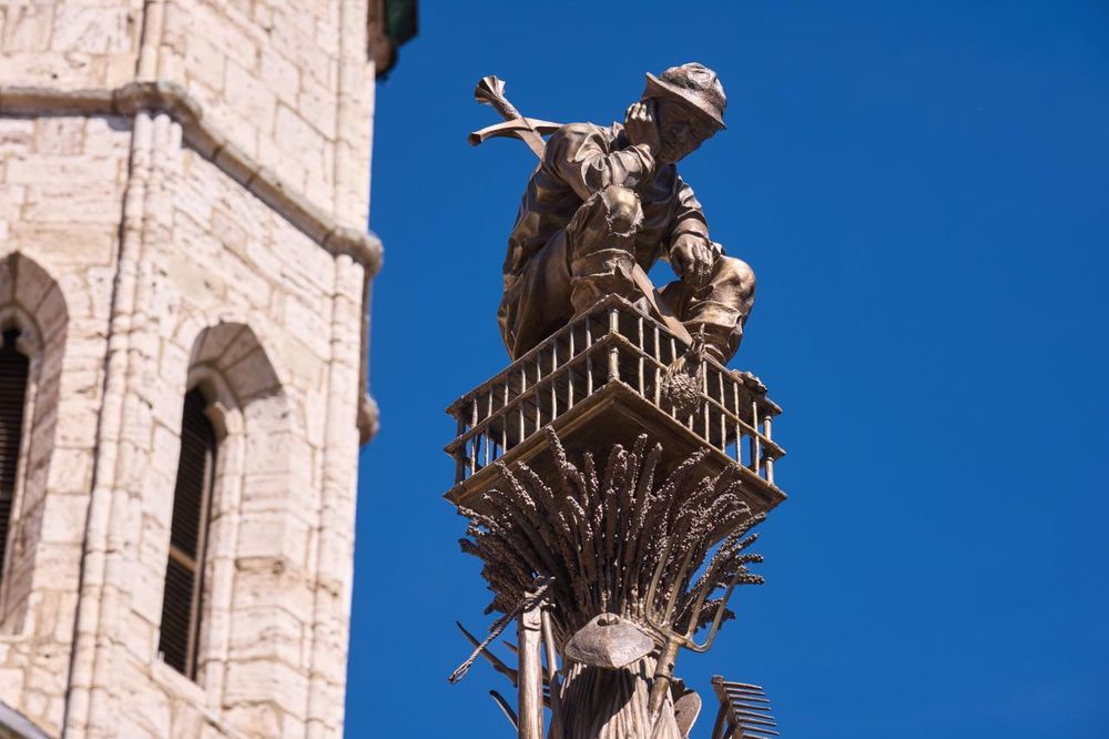 life size bronze statue of a despondent peasant seated head in hand on a chicken cage, a large sword protruding from his back, seen from below and silhouetted against blue sky, with a pale stone church tower to the left.