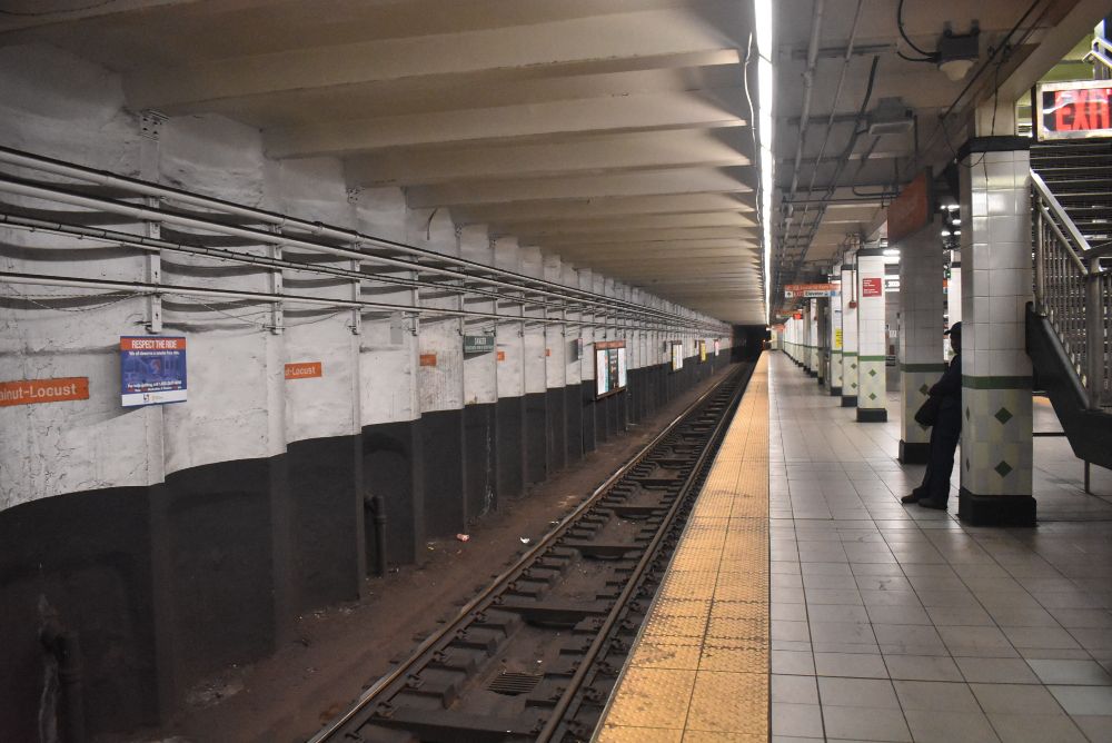 Walnut-Locust subway station. Looking south. wall with pipes and signage on left, tracks at bottom left, platform with exit sign and stairs on right of picture.