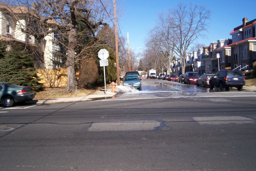 Picture of running fire hydrant on corner of 6th street and 66th avenue looking down 6th street with 66th avenue in foreground. A blue car sit behind the hydrant.