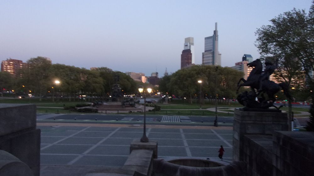 Partial skyline of Philadelphia from the museum of art. Trees in the left background, buildings and trees in the right background alongside the parkway, George Washington statue in the middle, and the courtyard and water fountain in the foreground.
