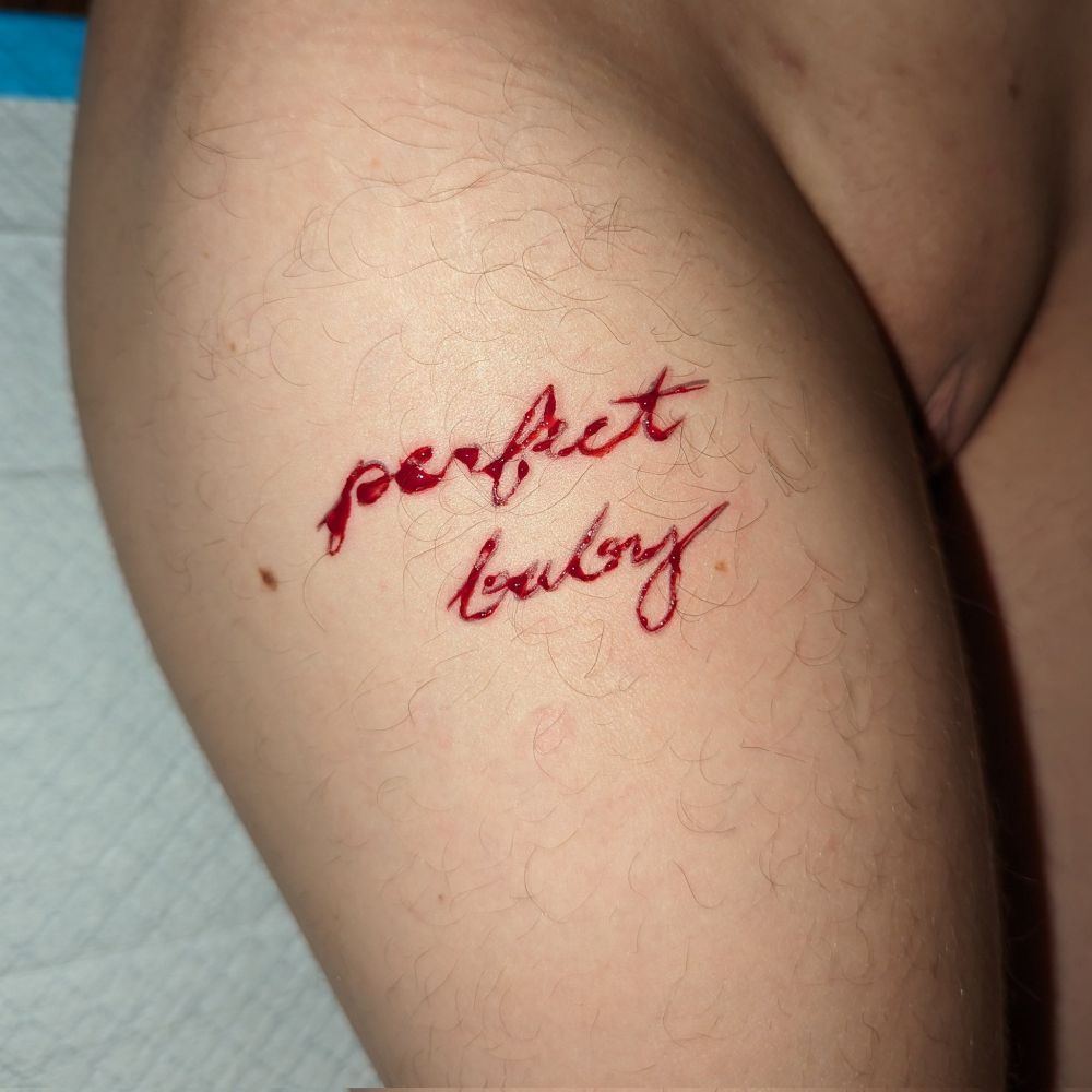 Square-cropped, flash photo of "perfect baby" carved in cursive, one word above the other, on Kit's outer thigh. He's naked and you can just see the root of his sensitive t-dick tucked between his legs. There's a puppy pad under his ass. He looks so soft and vulnerable, and he must have held so perfectly still during this carving. What an angel. Carta is lucky.
