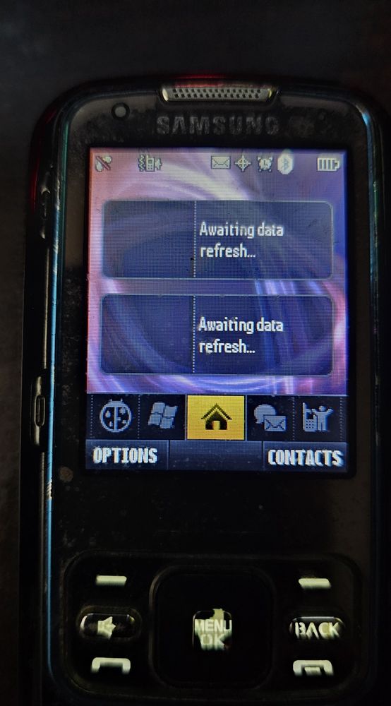 A Samsung Rant turned on. The display shows the home screen. Both widgets displayed say "Awaiting data refresh..." There is a menu bar at the bottom with five icons displayed: WeatherBug, the Windows logo, a home icon, messaging, and an icon of a person and a cell phone. Below the menu bar are two options selectable using two buttons on the face of the phone: "OPTIONS" and "CONTACTS".