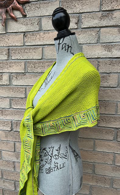 A headless mannequin wears a bright yellow shawl with a hint of a green tone. The edging of the shawl is done as a row of mitred squares in the yellow and a variegating yarn with pastels and greys. The same contrast colour runs in a single garter stitch ridge around the neck edge.