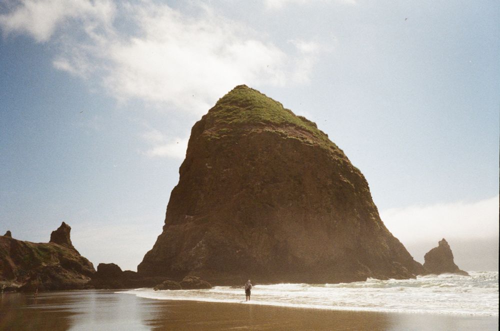 A film photograph of haystack rock which is a big rock just off the shore of cannon beach. One person is standing in the center of the frame in the surf with their back to the camera looking at the rock 