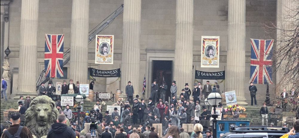 A crowd of people on the steps of St. George's Hall in Liverpool dressed in 19th Century clothes during filming for the upcoming Netflix series House of Guinness. There are Union flag banners and 