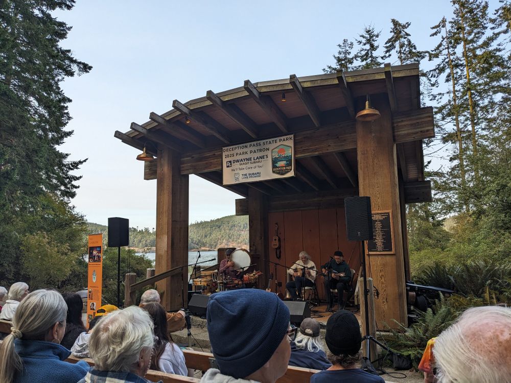 The image depicts an outdoor musical performance at Deception Pass State Park.  A three-person band is playing on a small stage under a wooden shelter. The band consists of musicians playing a multiple guitars, drums, and other string instruments. An audience is seated in front of the stage, enjoying the performance. The setting is scenic, with water visible in the background, and evergreen trees surrounding the performance area. The overall mood is peaceful and idyllic, suggesting a community gathering focused on music and nature. 