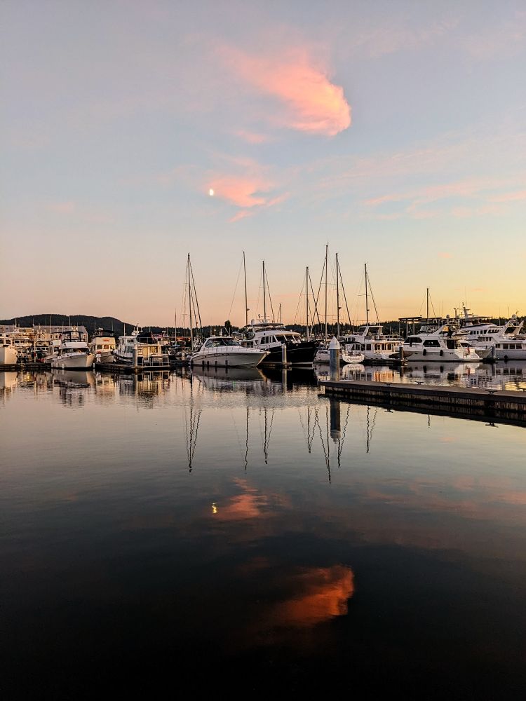 A tranquil marina scene at sunset or sunrise. A calm body of water reflects a pastel sky with hints of pink and peach. The clouds are soft and wispy, and a crescent moon is visible. Numerous sailboats and motor yachts are moored in the marina. The vessels are predominantly white with darker masts and rigging. The water's surface mirrors the sky and boats, creating a serene image. There's a half moon in the sky and one in the water. 