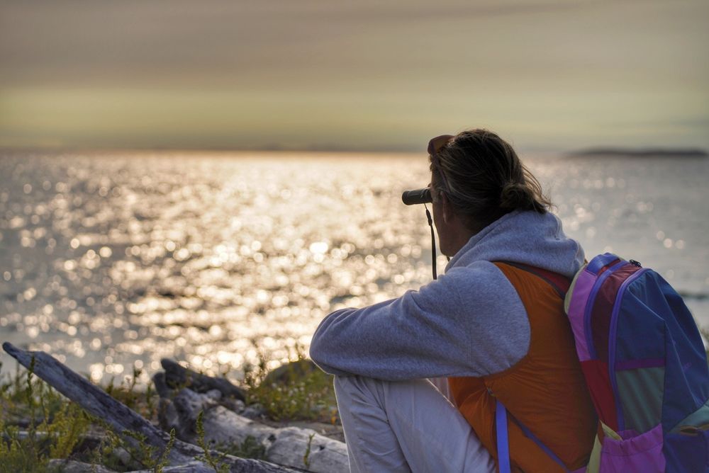 The image shows a man sitting on a rocky shore, looking out at a sparkling body of water at sunset. He is wearing a grey sweatshirt, light-colored pants, an orange vest, and a colorful backpack. He is holding binoculars to his eyes, seemingly observing the scene before them. The overall mood is peaceful and contemplative. The background features a calm sea reflecting the golden hues of the sunset, creating a bokeh effect. The foreground is rocky and shows some sparse vegetation.