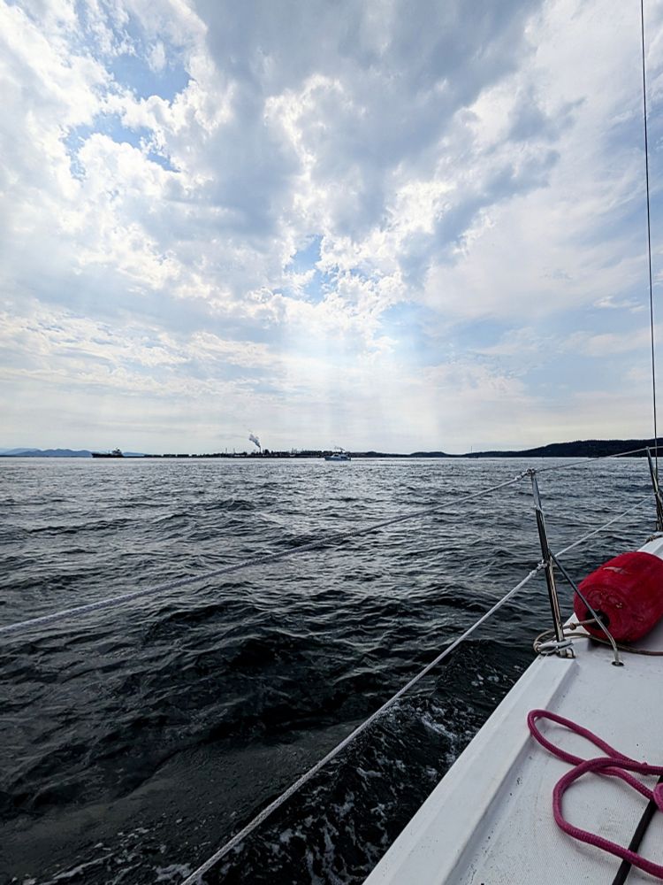 Full outdoor shot of a body of water from a sailboat under a partly cloudy sky. The water is a dark gray-blue, with visible wave patterns. A sailboat's deck, white in color, is at the bottom of the image, extending diagonally across the frame. Visible on the deck are ropes, and a red cylindrical fender is partially visible on the right side. The sky is mostly covered with puffy white and gray clouds, with some areas of lighter blue sky.  A few rays of light seem to be breaking through the clouds. In the distance, on the horizon, there's a glimpse of islands. 