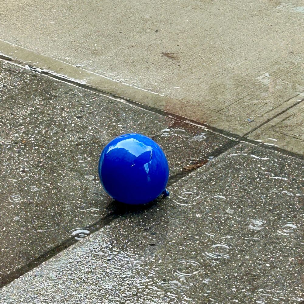 a shiny round blue balloon sites on a sidewalk in the rain