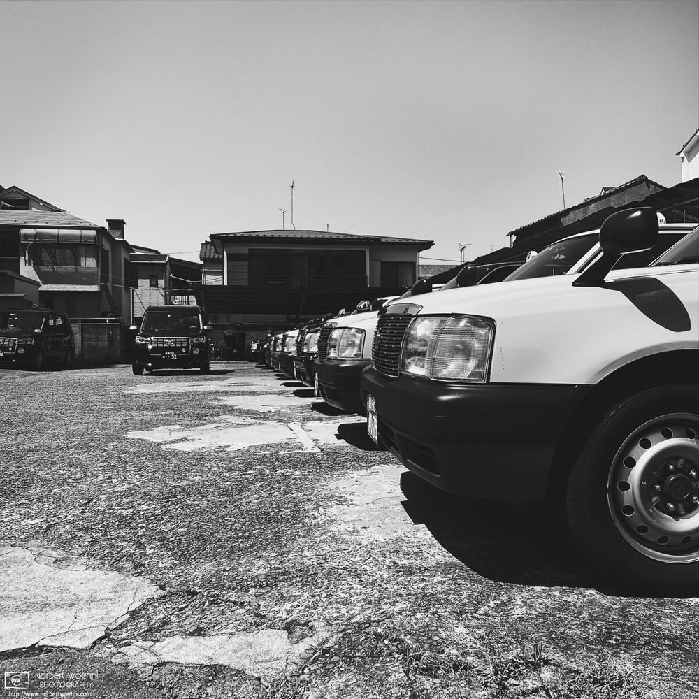 High-contrast monochrome photograph of a line-up of old taxis parked at "Tohto Taxi" in the Azusawa neighborhood of Itabashi Ward in Tokyo, Japan.