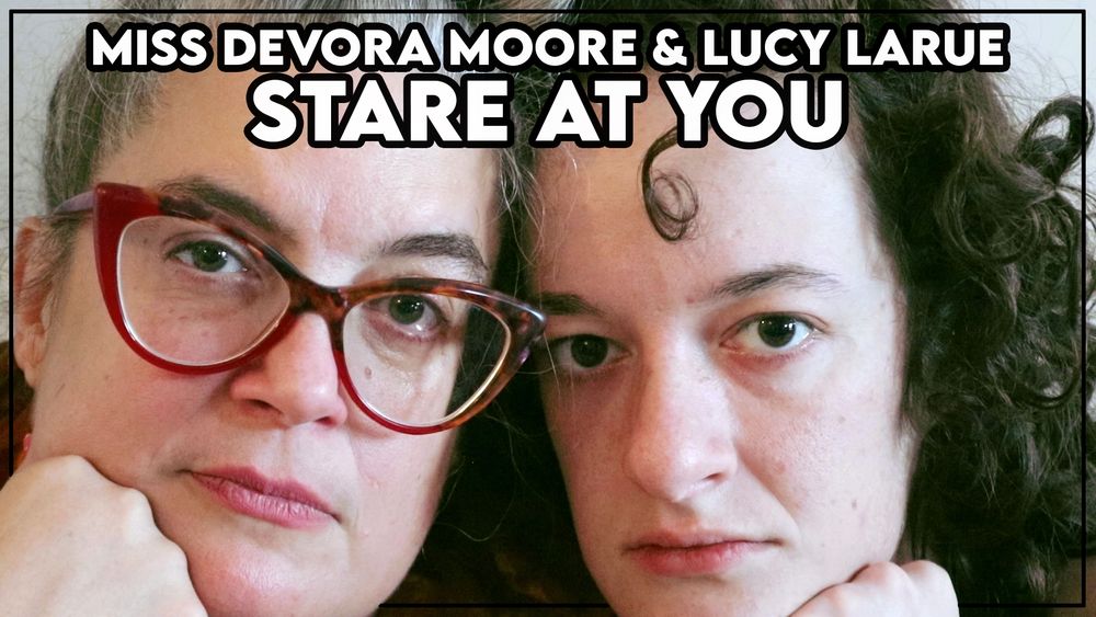 A title card for an age gap lesbian staring fetish femdom video called “Miss Devora Moore and Lucy LaRue Stare at You” showing two women's faces filling the screen, positioned very close to each other as they stare at you with blank expressions. On the left is Miss Devora Moore, Octogoddess, with turtled cat eye red glasses, blue-green eyes, and pink lipstick, her grey hair up in a bun, and on the right is Lucy LaRue, with curly brown hair and brown eyes, both with chins resting on their hands to watch you.