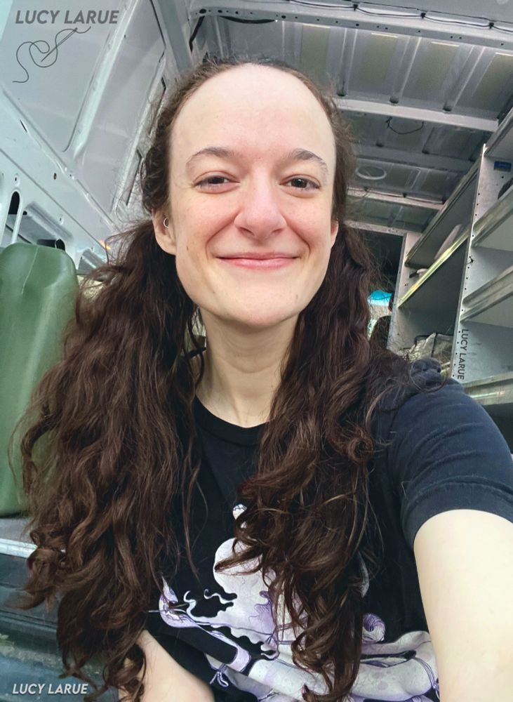 A smiling selfie taken by Lucy LaRue in he back of her sprinter van with shelving and a green water tank visible in the background. Lucy has her long curly brown hair in pigtails, is wearing a black shirt with hentai on it, and has a little ring piercing visible in her daith (ear).