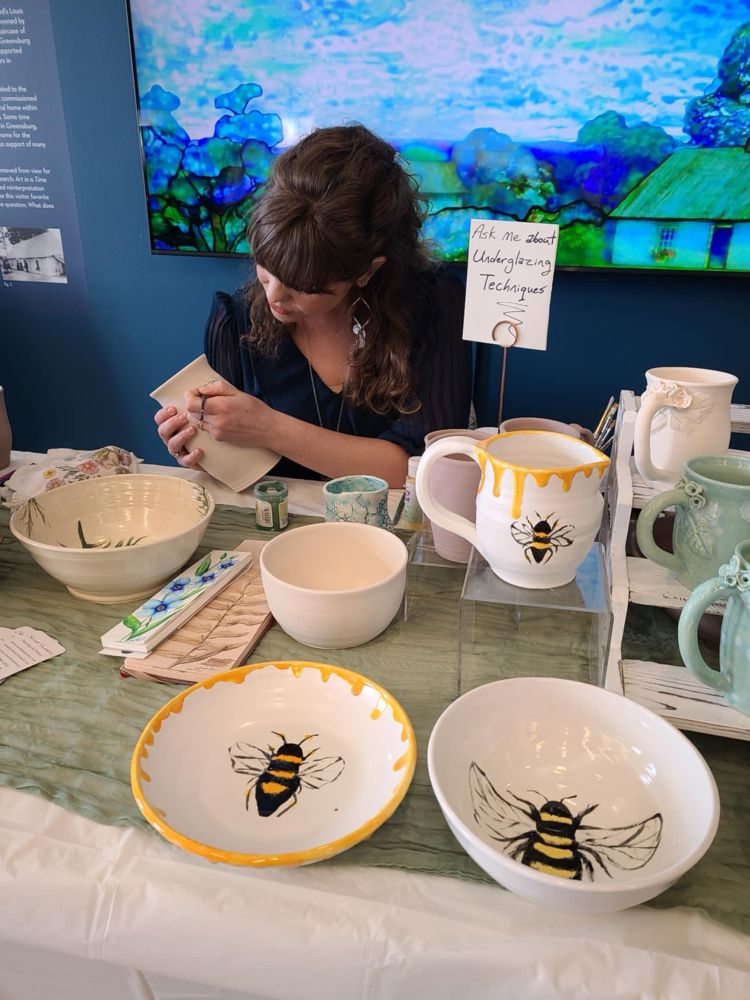A close up of a female artist painting small details on a vase. The details are of greenery. Several pieces of pottery are on the table in front of her.