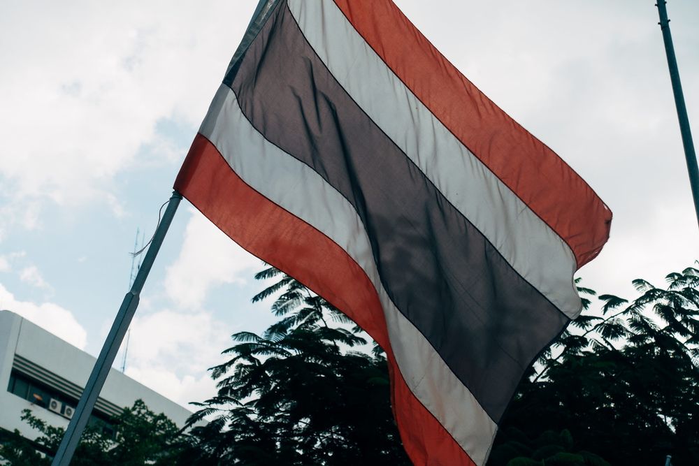 The flag of Thailand flying from the side of a building, with some greenery in the background.