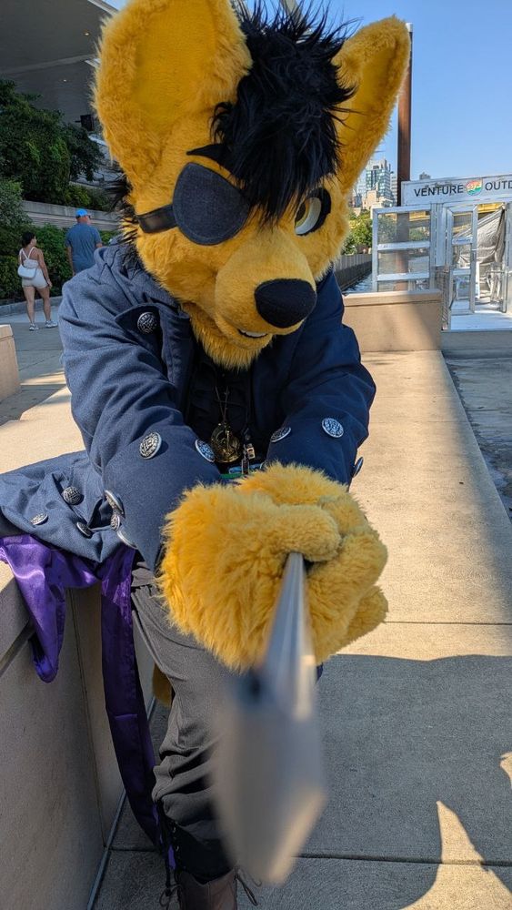 Fursuit photo of Caine, a yellow coyote in a black frilled pirate costume and a navy coat and purple sash

Lunging towards the camera the angle of the shot down the length of the sword