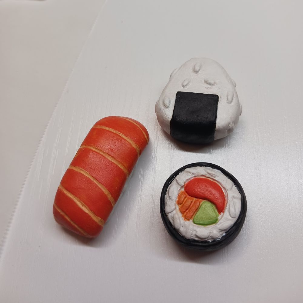 Three painted sushi made of polymer clay placed on a desk. There is a Salmon Nigiri, Onigiri, and a Maki Roll.