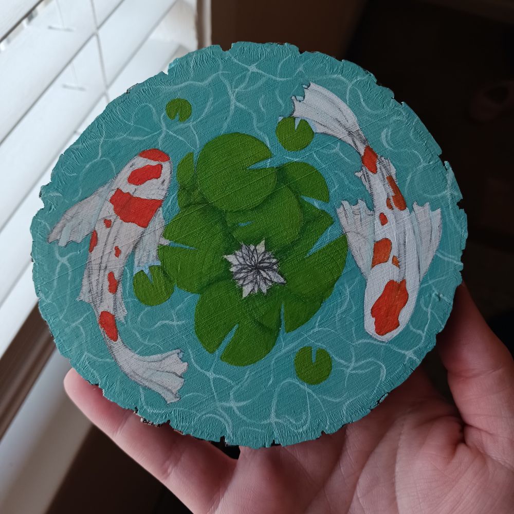 A work in progress painted koi pond on a round cut of wood. There are a gathering of green Lily pads and an unpainted flower in the center. Two orange spotted white koi with short flowy fins circling them while sun rays dance through the water.