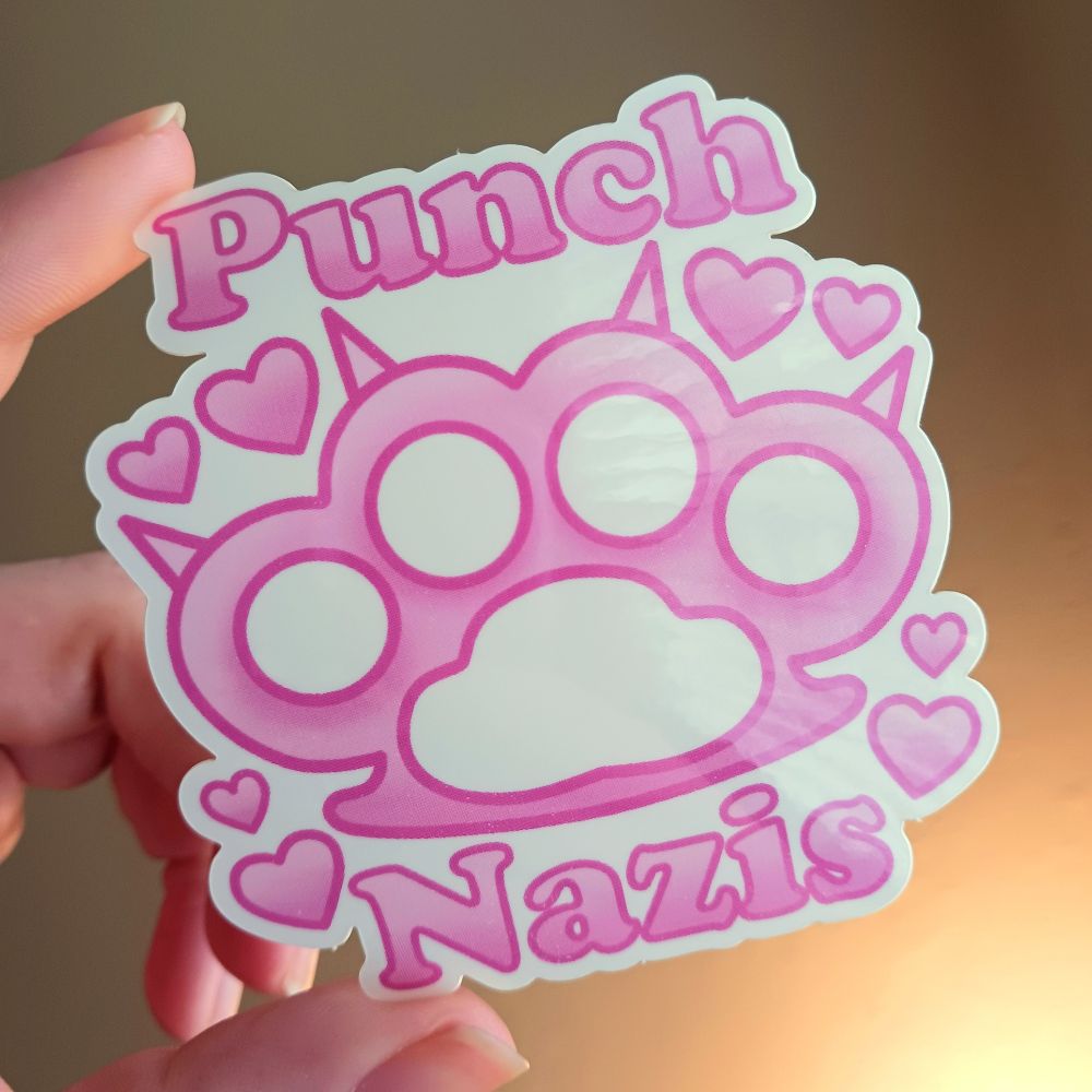 A "Punch Nazis" sticker being held by my hand, there are pink clawed brass knuckle with "Punch" in pink above them and "Nazis" in pink below with pink hearts around the brass knuckle, the background of the sticker is white.
