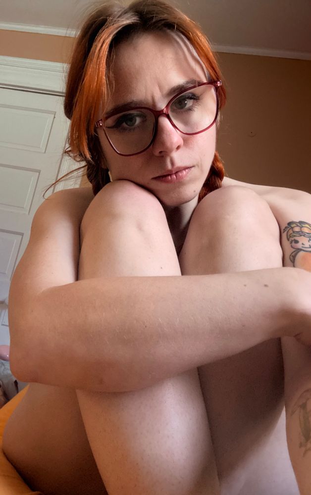 A woman with implied nudity sits with her legs crossed in front of her body.  she has on red square glasses and her hair is red in pigtails