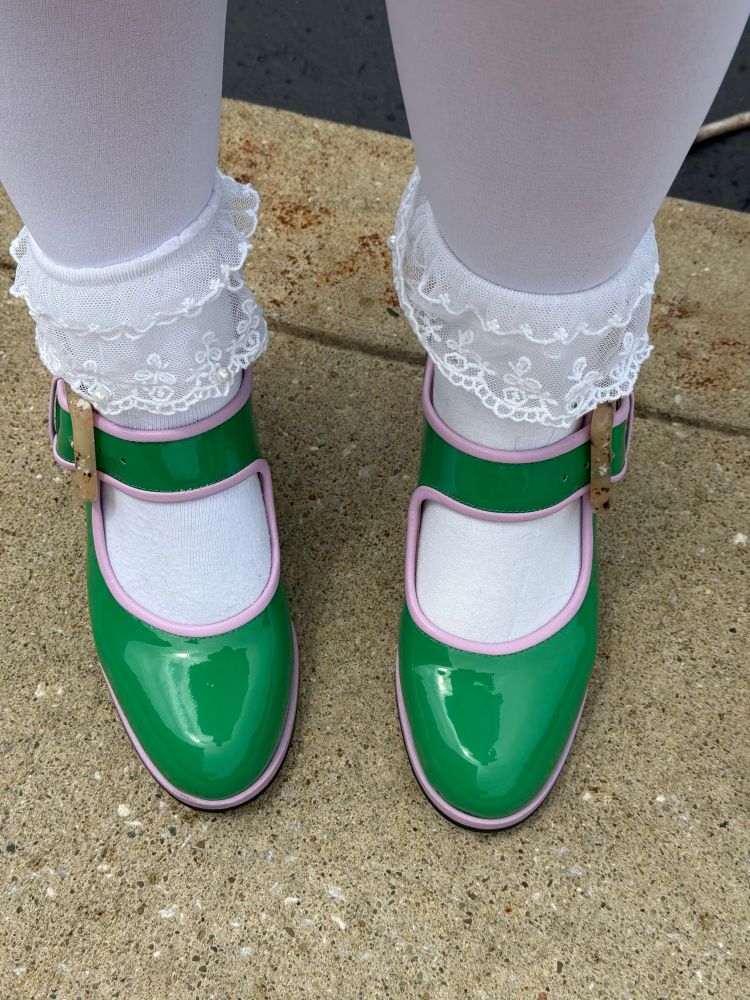 Green patent leather Mary Jane style shoes with lavender piping around the edges. They have a tortoise shell belt, buckle. The socks are white and Lacey with little frills around the ankle.
