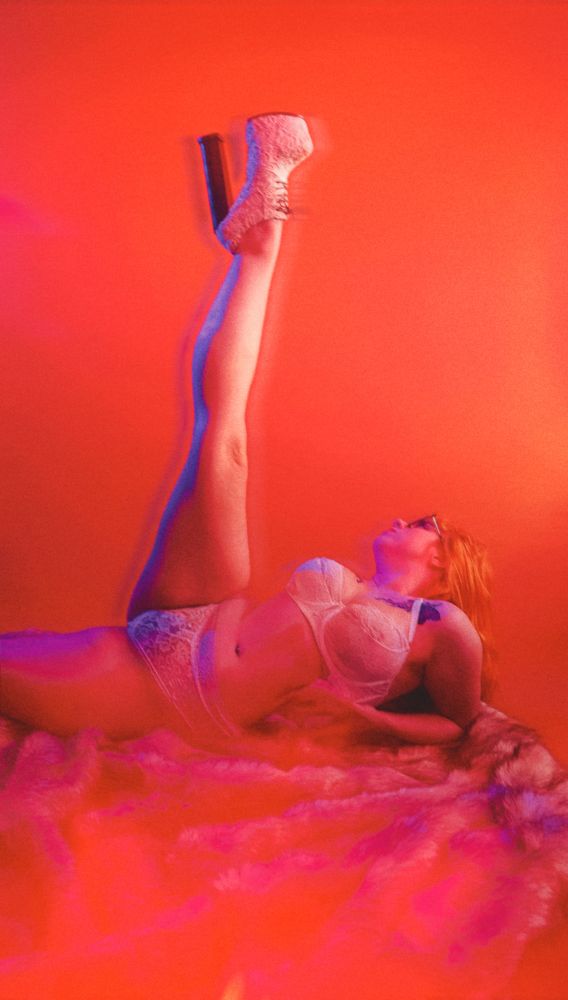 A woman with red hair and glasses in Lacey white lingerie lays on a furry blanket. She’s reclining on her back and stretching her leg up to the ceiling