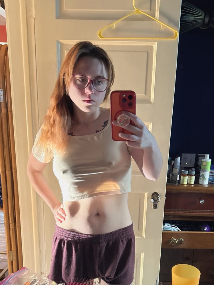 A redheaded woman taking a selfie in the bathroom mirror she wearing red glasses, maroon camp shorts, and a tan crop top. You can see the outlines of her nipples clearly through the crop top and her midriff is exposed with a soft belly.