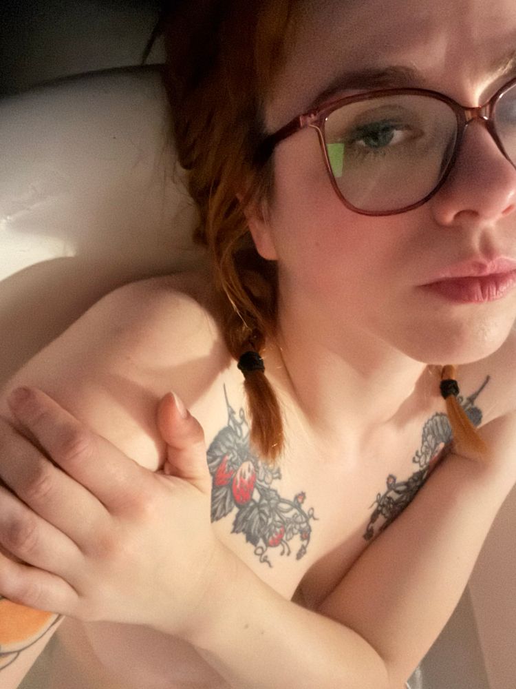 A woman with a chest tattoo, sleepy eyes, messy makeup and red hair in pigtails. She’s sitting in a bathtub