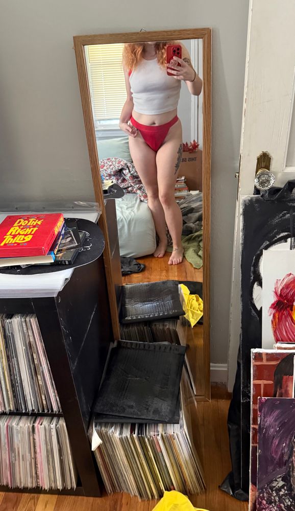 A woman’s lower torso and legs in the mirror. She has on a white crop top and he midriff is exposed. She’s not wearing pants and you can see her strappy red panties 