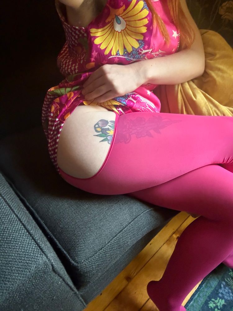 A a woman in a pink floral dress, sitting on a blue chair, lifts up her skirt to reveal a tattoo of a knife and magenta colored thigh high nylons