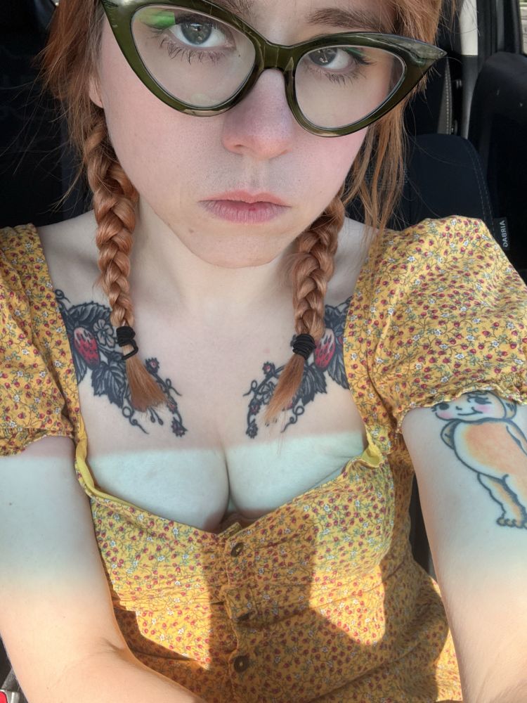 A woman with pale skin and red hair, pulled back in pigtails, stares up at the camera with big blue eyes from behind cat eye glasses. She’s wearing a floral yellow dress that is low-cut and you can see her cleavage, and that on her chest She has a strawberry tattoo.