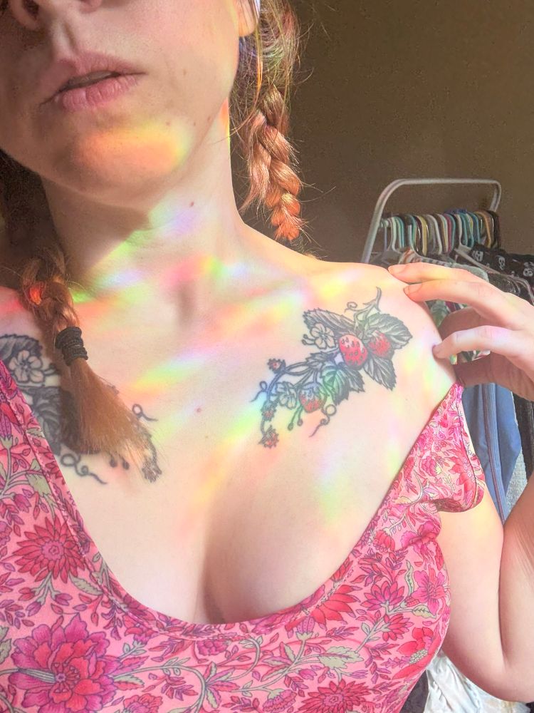 A woman with her red hair and pigtails, pulls down the shoulder of her pink Paisley dress to expose her clavicle and a chest tattoo of strawberries. You can only see her lips on her face, but there’s definitely some cleavage peeking out below