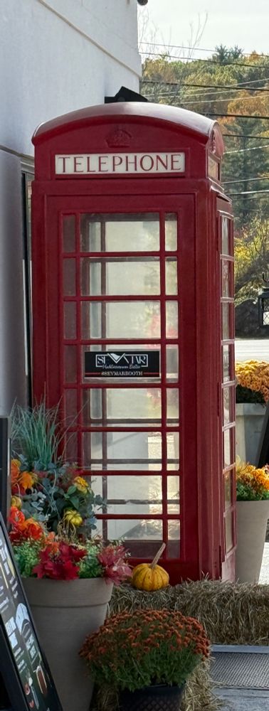 A red replica telephone booth with flowers and gourds all around with fall foliage in the background 