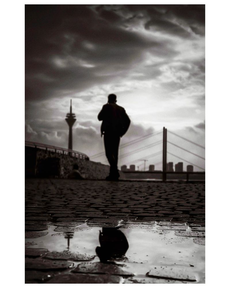 S/w Foto nach einem Regenschauer in der Düsseldorfer Altstadt..Mann,mit Handy am Ohr, spiegelt sich in in einer Pfütze und im Hintergrund sieht man den Rheinturm nebst Brücke ..Der Mann ist nur eine Silhouette 
