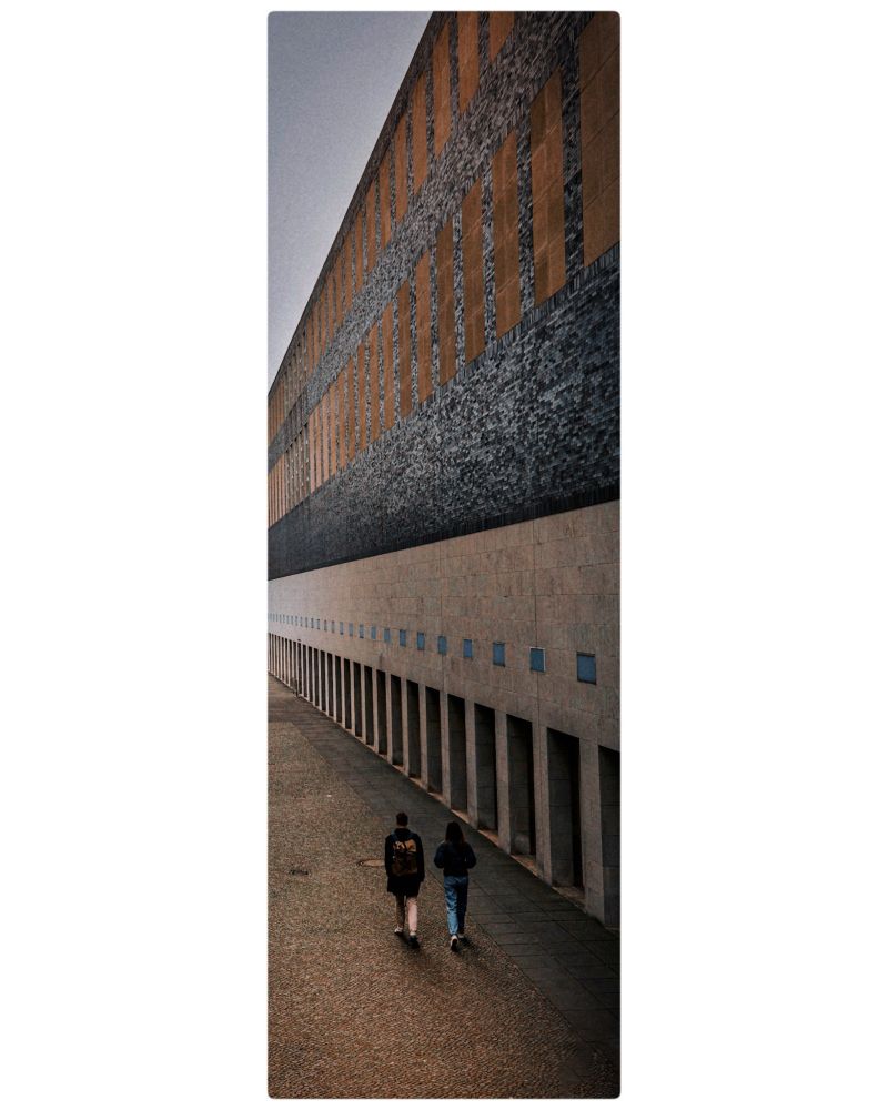 Rechts im Bild ist einen sehr hohe Mauer mit verschiedenen geometrischen Mustern.Unten,mittig im Bild sind zwei Personen die sich weg bewegen...Das Bild ist im Hochgebirge 
