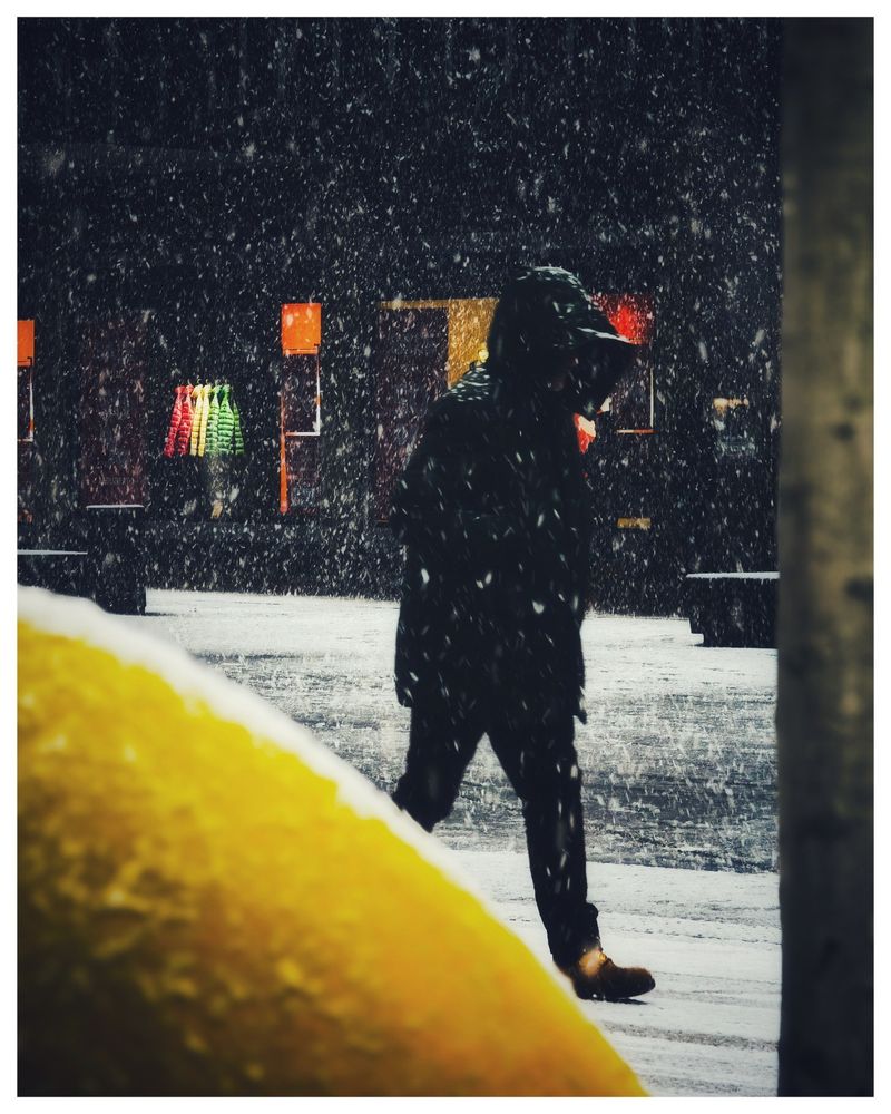 Eine dunkel gekleidete Person ,mit Kapuze,stampft in der Innenstadt durch den Schnee...Im Vordergrund sieht man eine Ecke von einem gelben Element 
