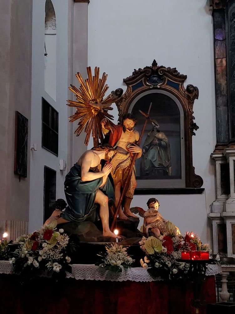 탁한 연분홍색 칠, 초록색 대리석 기둥, 성상을 모신 금색 테두리의 벽감으로 꾸민 흰 벽을 배경으로 흰 레이스 천을 덮은 붉은 벨벳재단 위에 꽃과 촛불, 아기천사들이 발치를 장식하는 예수와 십자가를 껴안고 그를 세례 중인 조반니 바티스타, 그들 뒤의 황금색 불꽃같은 장식이 보인다