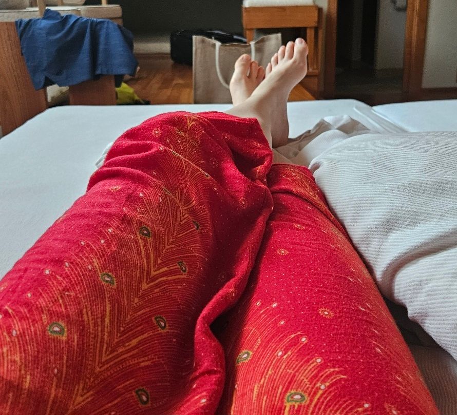 Das Bild zeigt eine Person, die auf einem Hotelbett liegt. Die Beine sind aus Perspektive der Person fotografiert. Man sieht sie ab den Oberschenkeln abwärts. Die Person trägt eine lange, rote Hose mit einem orange-gelben Pfauenfeder-Muster. Die Beine sind gekreuzt. Das Licht im Raum wirkt weich und diffus.
