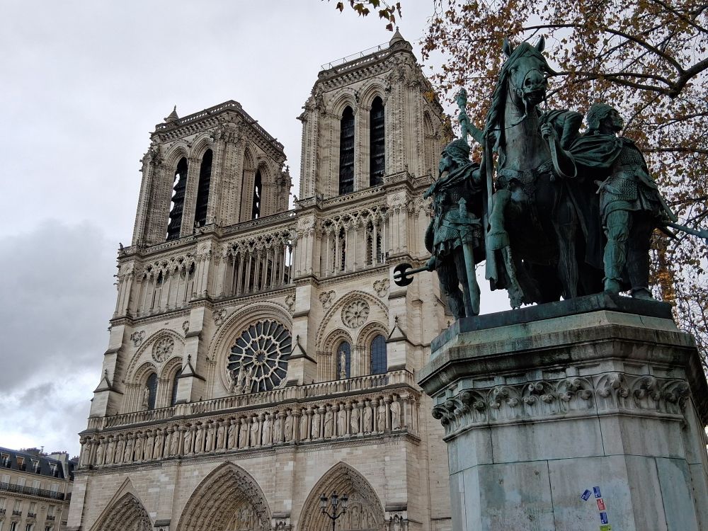 Façade de la cathédrale Notre-Dame sur fond de ciel gris ; au premier plan à droite, une statue équestre de Charlemagne.